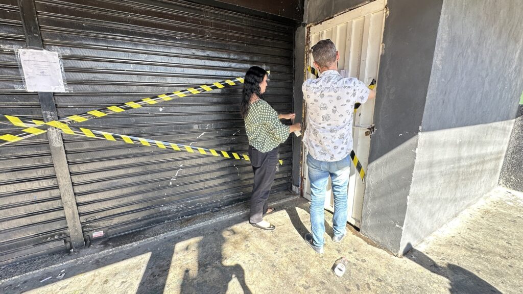 A imagem mostra a fachada de um estabelecimento comercial, possivelmente um restaurante, com a palavra "RESTAURANTE" e "INTERNACIONAL" visíveis na parte superior. A porta está fechada com uma grade metálica preta, e há uma fita de isolamento amarela e preta esticada na frente da entrada, indicando que o local está interditado ou fechado temporariamente. Na frente da porta, há duas pessoas: uma mulher de cabelos longos e escuros, vestindo uma blusa verde com padrão e calça preta, e um homem de cabelos grisalhos, usando uma camisa branca de manga curta com padrão e calça jeans. A mulher está segurando ou mexendo na fita de isolamento, enquanto o homem está próximo à porta branca ao lado da grade preta. No chão, próximo ao homem, há uma garrafa plástica descartada. A iluminação forte sugere que é dia, com sombras bem definidas no chão. A cena transmite a sensação de que o estabelecimento está fechado por algum motivo oficial ou de segurança.