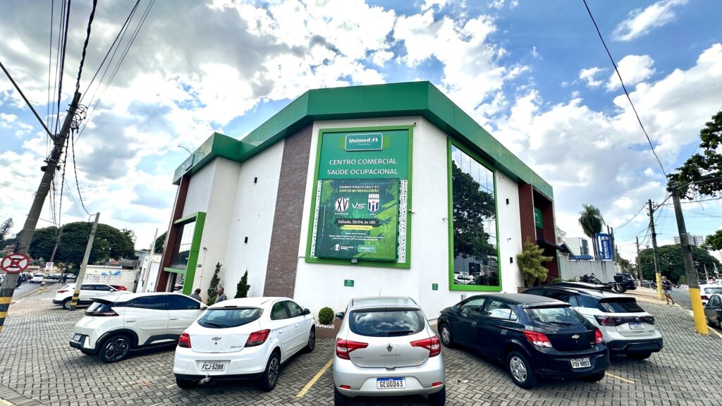 A imagem mostra um centro comercial moderno com fachada verde e branca, localizado em uma área urbana. Em frente ao prédio, há um estacionamento com vários carros estacionados, de diferentes cores, como branco, prata e preto. O céu está parcialmente nublado, com nuvens volumosas e brancas, contrastando com o azul do céu, indicando que é durante o dia. Na fachada do edifício, há uma grande placa verde com informações em português, que menciona "CENTRO COMERCIAL", "SAÚDE OCUPACIONAL" e "UNIMED PIRACICABA E XV". Também há uma referência a um evento esportivo, especificamente um jogo de futebol que acontecerá no sábado, 25 de abril, às 18h, com a frase "JUNTOS NO BRASILEIRÃO". A cena transmite um ambiente urbano tranquilo, com infraestrutura moderna e um clima agradável, sugerindo uma área de serviços e comércio. Ao fundo, é possível ver postes de energia e algumas árvores, complementando o cenário.