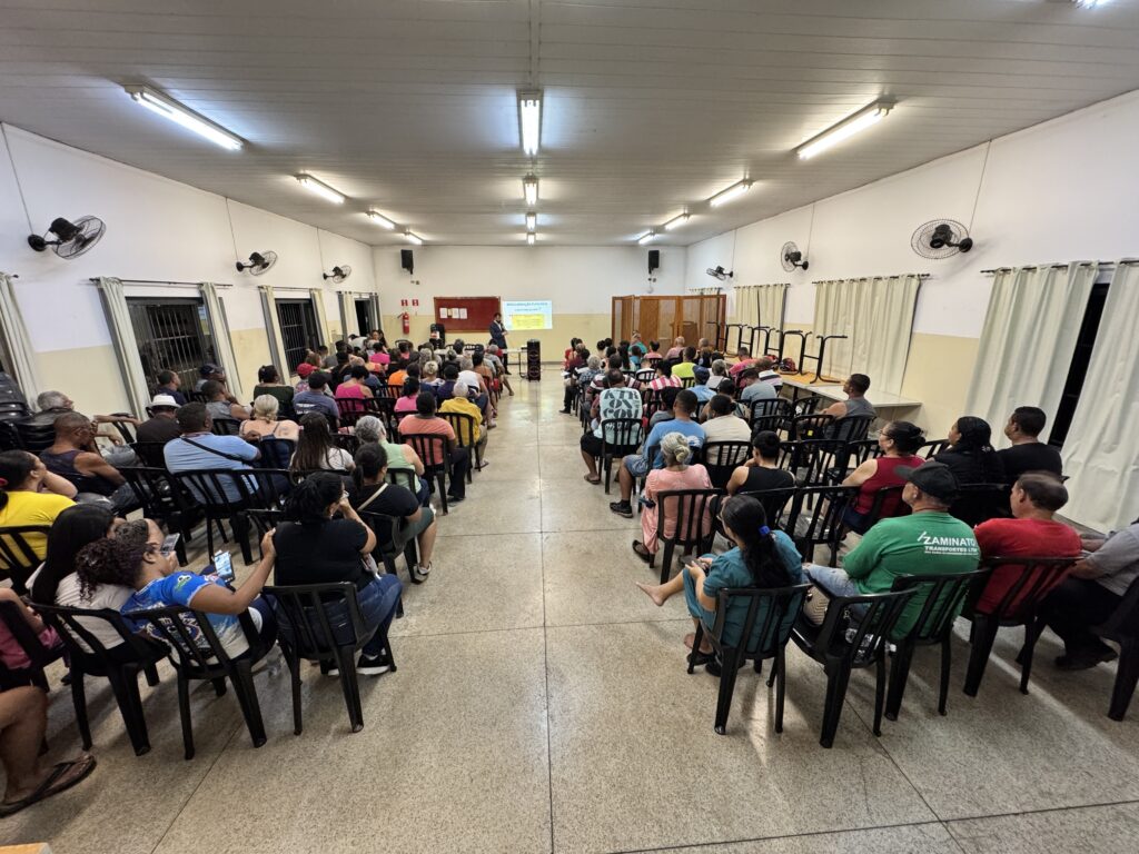 A imagem mostra uma reunião comunitária realizada em uma sala ampla e bem iluminada. A perspectiva é central, com um corredor no meio que divide fileiras de cadeiras pretas onde dezenas de pessoas estão sentadas, todas de costas para a câmera. A maioria dos participantes parece estar atenta à apresentação que ocorre na frente da sala, onde um homem está em pé próximo a uma tela de projeção. Na tela, lê-se o título "REGULARIZAÇÃO FUNDIÁRIA - O que é e como funciona?", indicando que o tema da reunião é sobre regularização fundiária. Ao lado da tela há um painel vermelho, possivelmente com informações adicionais. As paredes são brancas e há várias janelas com cortinas claras, além de ventiladores de parede e luminárias fluorescentes no teto, proporcionando uma iluminação uniforme. O ambiente sugere um encontro formal voltado para a comunidade, com o objetivo de informar ou discutir questões relacionadas à regularização de propriedades ou terrenos.