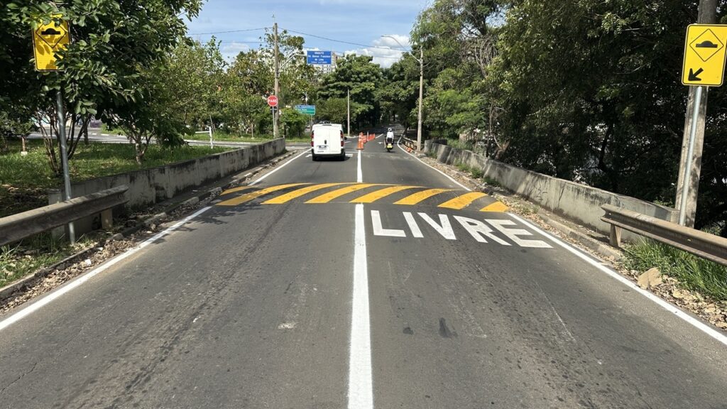 A imagem mostra uma estrada de duas pistas, cercada por árvores verdes de ambos os lados, sugerindo um ambiente tropical ou subtropical. No centro da pista, há um quebra-molas pintado com listras amarelas e brancas, indicando aos motoristas que reduzam a velocidade. Logo após o quebra-molas, está pintada no asfalto a palavra "LIVRE", em letras grandes e brancas, sugerindo que o trecho está livre ou desobstruído. Do lado esquerdo da estrada, há uma placa amarela em formato de losango que alerta sobre a presença do quebra-molas, representado por um símbolo preto de um carro passando sobre um ressalto. Do lado direito, outra placa amarela com uma seta preta indica a direção do quebra-molas. Também há guard-rails metálicos e barreiras de concreto ao longo dos dois lados da estrada. Ao fundo, é possível ver uma van branca e uma pessoa em uma motocicleta ou scooter seguindo pela via. Há cones de trânsito posicionados na pista, indicando uma possível área de trabalho ou desvio. Placas adicionais, incluindo uma placa de "PARE" em vermelho, estão visíveis mais adiante. O céu está parcialmente nublado, e a iluminação indica que é durante o dia, com sombras bem definidas. A estrada apresenta algumas marcas de desgaste, como rachaduras e sujeira, mostrando que é uma via usada com frequência. No geral, a imagem transmite uma sensação de uma estrada em área verde, com sinalização clara para garantir a segurança dos motoristas e pedestres.