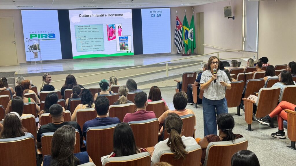 A imagem mostra uma palestra em um auditório, com o tema "Cultura Infantil e Consumo". A palestrante, uma mulher de cabelos curtos e óculos, veste uma camisa listrada branca e cinza e calça azul, está de pé ao lado dos assentos, segurando um microfone enquanto fala para a plateia. O público é composto principalmente por mulheres, sentadas em cadeiras de madeira, vistas de costas. No palco, há uma grande tela digital dividida em três partes. A parte central exibe o título da palestra e um texto relacionado à cultura infantil e consumo, mencionando conceitos como "Criança potencial consumidora" e "brinquedo sem caminho a percorrer", além de uma citação de Marcellino (1990). Também aparecem imagens das marcas Barbie e Minecraft, representando exemplos de consumo infantil. À esquerda da tela, há o logo da Prefeitura de Pira (possivelmente Piracicaba), e à direita, a data "31-03-2026" e o horário "08:59". Três bandeiras (do estado de São Paulo, do Brasil e do município) estão posicionadas ao lado direito do palco. O ambiente é um auditório bem iluminado, com paredes claras e cadeiras organizadas em fileiras, criando um cenário típico de evento acadêmico ou institucional.