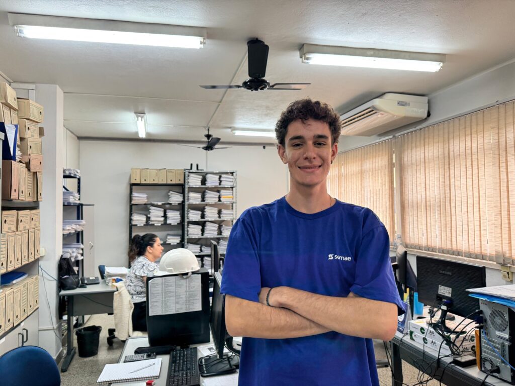 A imagem mostra um ambiente de escritório, com iluminação fluorescente e paredes claras. Em primeiro plano, há um jovem homem de cabelos cacheados castanhos, sorridente, vestindo uma camiseta azul com o logo "semae". Ele está de braços cruzados e olhando diretamente para a câmera. Atrás dele, à esquerda, uma mulher está sentada em uma mesa, aparentemente trabalhando, olhando para o lado. No escritório, há vários objetos visíveis, incluindo monitores de computador (um deles marca Samsung), teclados, uma calculadora, um capacete branco sobre a mesa, cadernos e pilhas de papéis. Nas prateleiras ao fundo, há muitas caixas de arquivo organizadas. As janelas têm persianas verticais que deixam entrar luz natural, indicando que é dia. O ambiente parece ser um escritório administrativo, possivelmente relacionado a serviços públicos, já que o "semae" normalmente está ligado a serviços de água e saneamento.