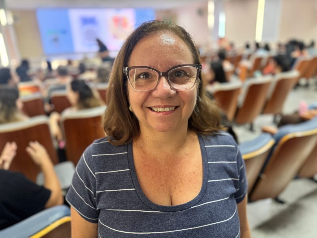 A imagem mostra uma mulher de meia-idade com cabelo castanho ondulado, na altura dos ombros, usando óculos de aro grosso e escuro. Ela está sorrindo de forma amigável e olha diretamente para a câmera. O fundo está desfocado, mas é possível ver várias pessoas sentadas em cadeiras, como em um auditório ou sala de conferências. A iluminação é clara e o ambiente parece ser interno, possivelmente durante um evento, palestra ou seminário. A mulher está em foco nítido, enquanto os demais elementos ao redor estão desfocados, destacando-a como o ponto principal da foto.
