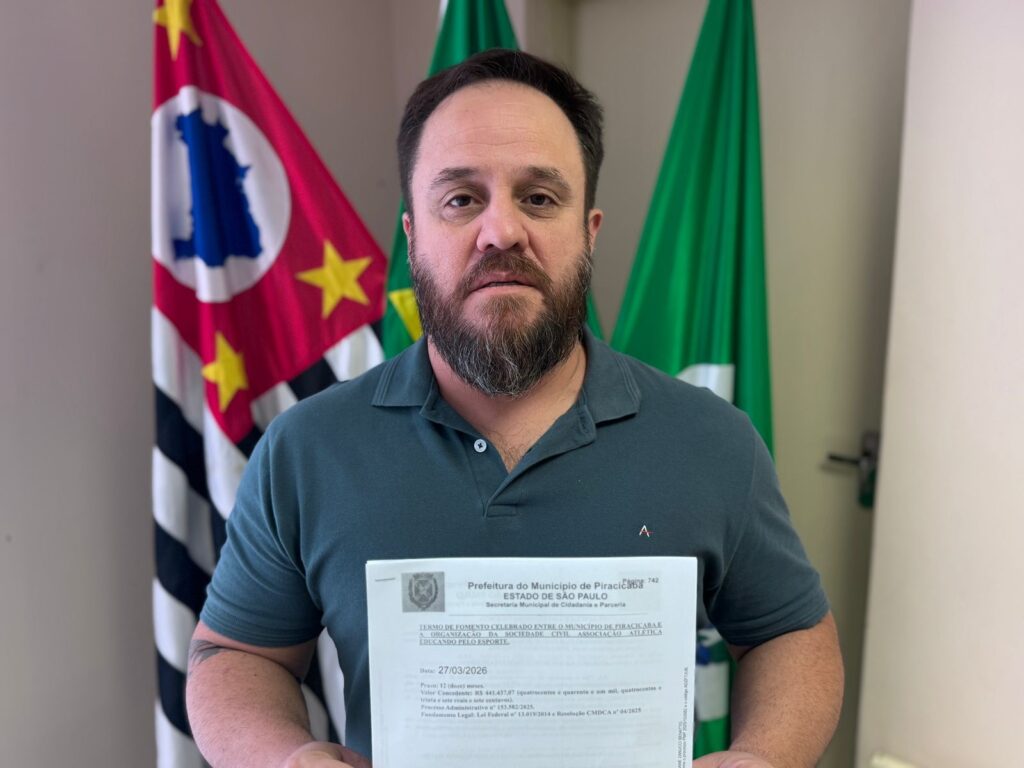 A imagem mostra um homem em pé, segurando um documento oficial. Ele está vestido com uma camisa polo verde escura e tem cabelos e barba escuros. Atrás dele, há duas bandeiras: uma com as cores vermelho, branco e amarelo, possivelmente a bandeira do município de Piracicaba, e outra bandeira verde. O homem está olhando diretamente para a câmera, com expressão neutra. O documento que ele segura é um "Termo de Fomento" celebrado entre o Município de Piracicaba, no Estado de São Paulo, e a organização da sociedade civil "Associação Atlética Educando pelo Esporte". O termo tem data de 27 de março de 2026, com prazo de 12 meses. O valor concedido é de R$ 441.437,07. Também são mencionados o número do processo administrativo e a fundamentação legal para o termo. A imagem parece representar a formalização de um convênio ou parceria entre a prefeitura e a associação mencionada, provavelmente para o desenvolvimento de atividades esportivas ou sociais.