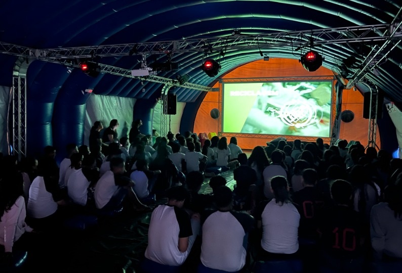 A imagem mostra um evento acontecendo dentro de uma grande tenda inflável azul, com um teto curvo sustentado por estruturas metálicas. No centro, há uma grande tela exibindo o nome "MICHAEL ADAMS" em letras brancas, com um fundo em tons de verde e laranja. A frente da tela está cheia de pessoas, principalmente jovens, sentadas no chão e assistindo à apresentação. A iluminação é baixa, com luzes focadas na tela e algumas luzes vermelhas criando um efeito dramático no ambiente. A tenda parece ser um espaço temporário, provavelmente usado para palestras, apresentações ou eventos culturais. A atmosfera é de concentração e interesse por parte do público. As cores predominantes são o azul da tenda e o contraste das luzes laranja e vermelha, dando um visual vibrante ao espaço.