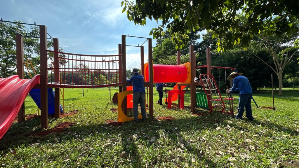 A imagem mostra um parquinho infantil em processo de montagem ou instalação em um ambiente externo, provavelmente um parque. O brinquedo principal é uma estrutura grande e colorida, composta por escorregadores vermelhos, uma ponte ou passarela vermelha, uma rede para escalar e um escorregador em formato de túnel laranja. Dois homens estão trabalhando na montagem: um está próximo aos escorregadores e o outro está usando uma pá próximo à rede de escalada. O chão ao redor da estrutura está mexido, com terra exposta, indicando que o parquinho está sendo instalado recentemente. O local é gramado, com algumas folhas espalhadas, e há árvores ao fundo com folhagem verde. O céu está azul com algumas nuvens brancas, indicando um dia ensolarado. A iluminação destaca as cores vibrantes do parquinho, e a foto foi tirada de um ângulo ligeiramente baixo, dando uma sensação de imponência à estrutura.