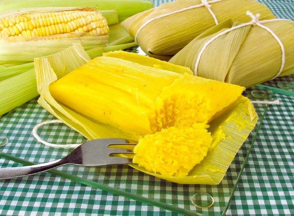 A imagem mostra uma foto em close de vários tamales, um prato tradicional latino-americano, dispostos sobre uma toalha de mesa xadrez verde e branca. Os tamales estão envoltos em folhas de milho, com alguns parcialmente abertos, revelando o recheio amarelo vibrante. Em primeiro plano, um tamal está aberto e um garfo está pegando um pedaço, indicando que está pronto para ser comido. Ao lado dos tamales, há uma espiga de milho ainda com a casca, remetendo ao ingrediente principal do prato. A iluminação é natural e clara, destacando as cores vivas do recheio e das folhas. O foco da foto está nos tamales, com o fundo desfocado para chamar atenção à comida, transmitindo uma sensação de comida caseira e apetitosa.