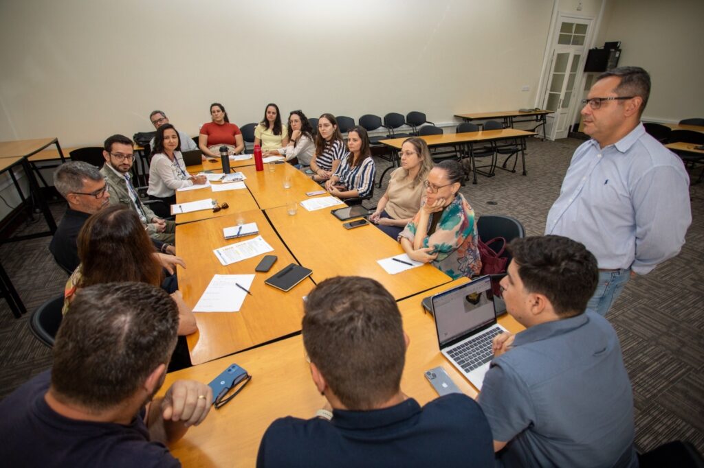 A imagem mostra uma reunião em uma sala de conferências, com várias pessoas sentadas ao redor de uma grande mesa de madeira polida. O ambiente é bem iluminado, com luzes no teto que proporcionam uma iluminação neutra. Sobre a mesa, há diversos objetos como papéis, canetas, laptops, celulares e garrafas de água, indicando que a reunião está em andamento e que os participantes estão engajados em discussões ou apresentações. Ao todo, são cerca de 12 a 15 pessoas, de diferentes idades e gêneros, vestindo roupas em estilo casual de negócios. Algumas pessoas estão concentradas em seus laptops ou documentos, enquanto outras conversam entre si. Um homem está em pé ao lado direito da imagem, aparentemente falando ou observando os demais. O fundo da sala tem cadeiras e mesas adicionais, sugerindo um espaço maior para reuniões ou aulas. A composição da imagem é uma visão média, capturando a mesa e os participantes, com uma perspectiva levemente elevada. As cores predominantes são os tons quentes da madeira da mesa, contrastando com as paredes e o tapete em tons neutros. A atmosfera geral é de colaboração e profissionalismo.