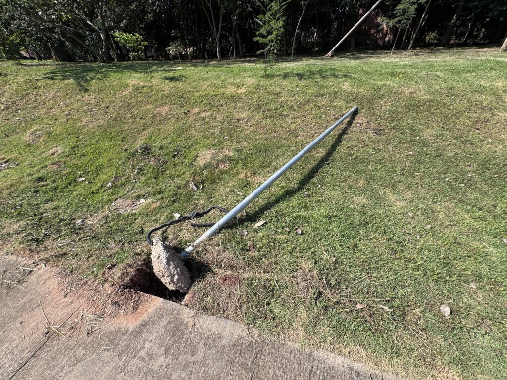 A imagem mostra um poste metálico caído no chão, em uma área gramada próxima a um caminho pavimentado. O poste, que parece ser de alumínio ou aço, está deitado sobre a grama, com sua base presa a um grande bloco de concreto que foi arrancado do solo, deixando um buraco visível na terra. Há fios elétricos pretos conectados à base do poste, que estão expostos. O gramado ao redor apresenta algumas manchas de grama seca e folhas espalhadas. Ao fundo, há uma fileira densa de árvores com folhagem escura, indicando que a área é provavelmente um parque ou uma área verde. A luz do dia ilumina a cena, criando sombras visíveis no chão. Não há pessoas na imagem, e o ambiente transmite uma sensação de abandono ou de um incidente recente que causou a queda do poste.