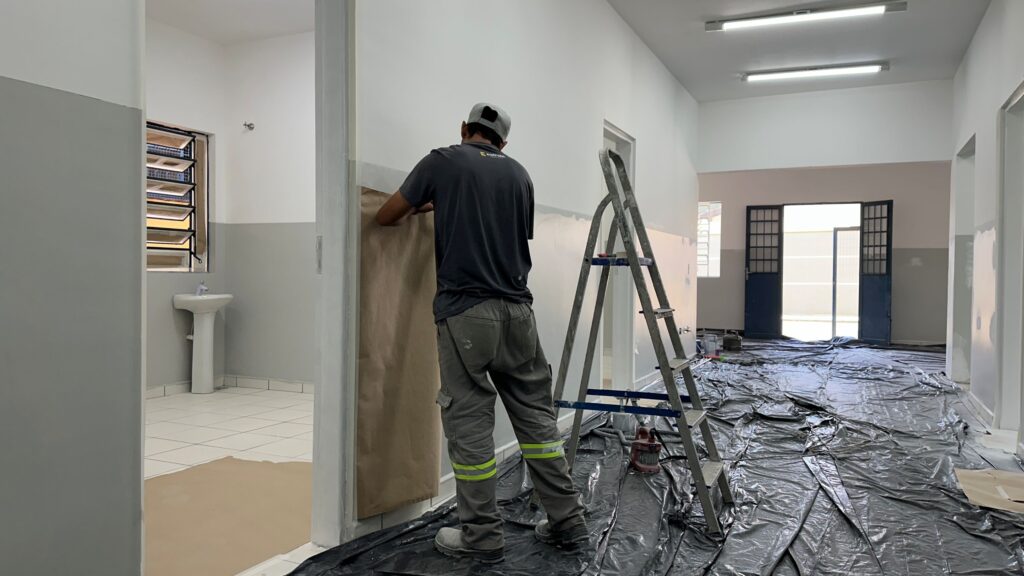 A imagem mostra um trabalhador em um ambiente interno, provavelmente um corredor de um prédio em reforma. Ele está de costas, vestindo camiseta cinza, calça de trabalho cinza com faixas refletivas amarelas e boné cinza. O trabalhador está aplicando um papel pardo adesivo na parede, possivelmente para proteger áreas durante a pintura. No chão, há um plástico preto cobrindo toda a superfície para proteger contra respingos. À direita do trabalhador, há uma escada de alumínio do tipo cavalete. Ao fundo, é possível ver uma pia branca e uma janela com venezianas. O corredor é iluminado por luzes fluorescentes no teto, e ao final do corredor há uma porta dupla azul. No verso da camiseta do trabalhador está escrito "estrela", que provavelmente é o nome da empresa ou equipe responsável pela obra. A cena transmite a ideia de um ambiente em processo de reforma ou pintura, com cuidados para proteger as superfícies.