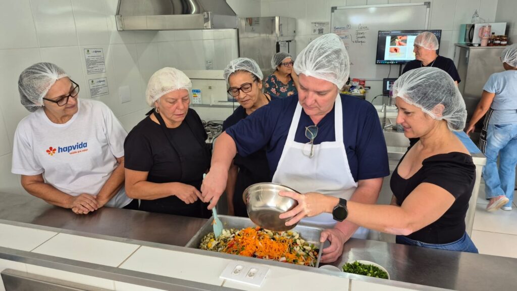 A imagem mostra um grupo de pessoas em uma cozinha, provavelmente participando de uma aula ou demonstração de culinária. Todos estão usando toucas para cabelo, indicando um ambiente que segue normas de higiene. No centro, uma pessoa está despejando ingredientes de uma tigela metálica para uma assadeira grande que contém legumes picados, como cenoura ralada. Ao redor, outras pessoas observam atentamente o processo. O ambiente é uma cozinha profissional ou educacional, com bancadas de aço inox, equipamentos e um quadro branco ao fundo com anotações e uma projeção, sugerindo que estão seguindo instruções ou uma receita. A atmosfera parece colaborativa e focada no aprendizado culinário.