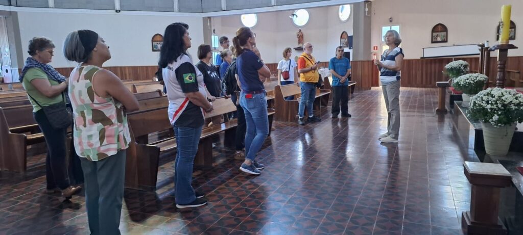 A imagem mostra um grupo de pessoas dentro de uma igreja. Elas estão reunidas em um corredor entre bancos de madeira, aparentemente ouvindo uma mulher que está falando, posicionada à direita da imagem. A mulher está vestindo uma blusa listrada e calças claras, e parece estar conduzindo uma visita ou explicação. O grupo é composto por cerca de dez adultos, todos vestidos de forma casual, com roupas como camisetas, calças jeans e coletes. Um dos participantes, em primeiro plano, veste uma camiseta branca com um patch da bandeira do Brasil na manga e as letras "SMO". O ambiente da igreja é iluminado por luz natural que entra por janelas circulares próximas ao teto. O chão é revestido por ladrilhos escuros com desenhos geométricos em tons avermelhados. À direita, próximo à mulher que fala, há um altar decorado com vasos grandes de flores brancas e uma vela amarela alta. As paredes são brancas e há elementos religiosos, como quadros pequenos e uma estátua, sugerindo que o local é tradicional e bem cuidado. A atmosfera parece ser de atenção e respeito, típica de uma visita guiada ou reunião em um espaço religioso.