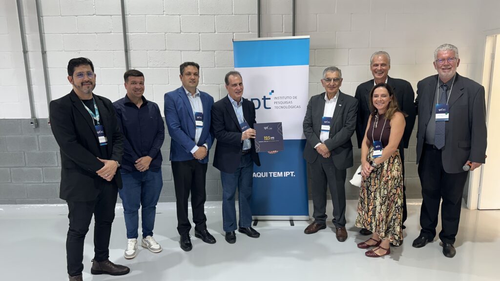 A imagem mostra um grupo de oito pessoas posando para uma foto em um ambiente interno, à frente de uma parede branca de tijolos. Entre elas, há sete homens e uma mulher. Alguns dos homens estão vestidos com ternos, enquanto outros usam roupas mais casuais, como camisa e calça jeans. A mulher está usando uma blusa sem mangas vinho e uma saia estampada longa. Uma das pessoas, no centro, segura um livro ou pasta azul escura com a inscrição "125 ANOS". Ao fundo, há uma faixa vertical azul e branca com o logotipo do "ipt INSTITUTO DE PESQUISAS TECNOLÓGICAS" e o slogan "AQUI TEM IPT". Também está escrito "125 ANOS" e "Construindo o futuro com Missão, Tecnologia e Inovação", indicando que a foto foi tirada em um evento comemorativo dos 125 anos do instituto. O ambiente parece moderno e bem iluminado, com um piso cinza claro e luzes industriais no teto. O clima da imagem é formal e profissional, sugerindo uma celebração ou reunião importante relacionada ao instituto.