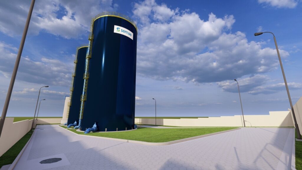 A imagem mostra uma instalação industrial, provavelmente uma estação de tratamento ou armazenamento de água. Em destaque, há dois grandes tanques cilíndricos de cor azul escura com tampas amarelas no topo. Cada tanque possui escadas externas e guarda-corpos amarelos para acesso e segurança. Na base dos tanques, é possível ver tubulações azuis conectadas, sugerindo o fluxo de líquidos. O ambiente ao redor é bem cuidado, com gramado verde ao redor dos tanques e um caminho pavimentado com blocos hexagonais em primeiro plano. A área está cercada por um muro baixo de cor clara, e há postes de iluminação distribuídos pelo local. O céu está azul com algumas nuvens brancas, indicando um dia ensolarado. A perspectiva da imagem é ligeiramente elevada, permitindo uma visão clara dos tanques e do entorno. A palavra "semae" está visível em um dos tanques, possivelmente indicando o nome da empresa ou órgão responsável pela instalação. A cena transmite uma sensação de organização, limpeza e modernidade na infraestrutura.