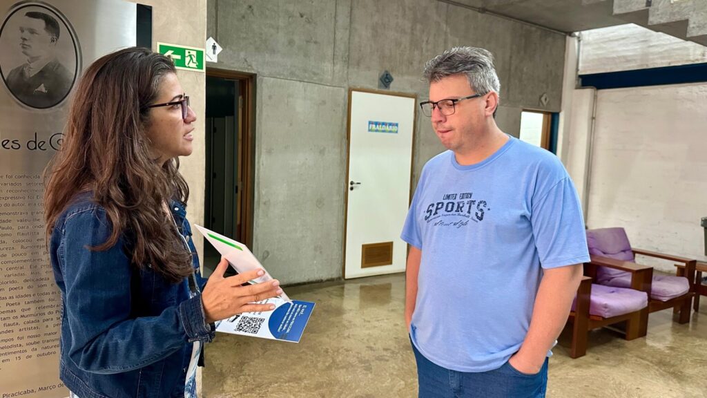 A imagem mostra duas pessoas conversando em um ambiente interno moderno. À esquerda, uma mulher com cabelo longo e escuro, usando óculos e uma jaqueta jeans, segura um folheto branco com um design azul e um código QR, enquanto fala e gesticula com a mão direita. À direita, um homem com cabelo curto e grisalho, também usando óculos, veste uma camiseta azul clara com a palavra "SPORTS" escrita e calça jeans, observando o folheto com as mãos nos bolsos. Ao fundo, há uma porta com a placa "FRALDÁRIO" e um pequeno cartaz azul, além de cadeiras roxas. O local apresenta paredes de concreto e um piso polido, com iluminação clara e uniforme. À esquerda, há uma placa memorial com um retrato e texto em português parcialmente visível. A cena parece ser em um espaço público, possivelmente uma instituição ou centro comunitário na cidade de Piracicaba, conforme indicado no folheto e na placa.