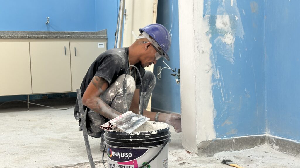 A imagem mostra um trabalhador da construção civil em um ambiente interno, aparentemente uma cozinha ou laboratório em reforma. Ele está agachado no chão, aplicando massa corrida ou algum tipo de revestimento branco em uma parede azul clara. O homem usa capacete azul, óculos de proteção transparentes, camiseta preta e calças que estão cobertas de pó branco, indicando o trabalho com materiais de construção. Seus braços também estão sujos com o mesmo pó. À sua frente, há um balde preto com a marca "UNIVERSO TINTAS" contendo o material branco que ele está usando, além de uma espátula metálica apoiada na borda do balde. A parede que ele está trabalhando tem uma parte já revestida com massa branca, enquanto outra parte ainda está azul. Ao fundo, há um armário com portas bege e uma bancada de granito com uma torneira, reforçando a ideia de que o espaço está em reforma. Também é possível ver fios elétricos expostos saindo de uma abertura na parede. A iluminação é clara e uniforme, típica de um ambiente interno durante o dia.