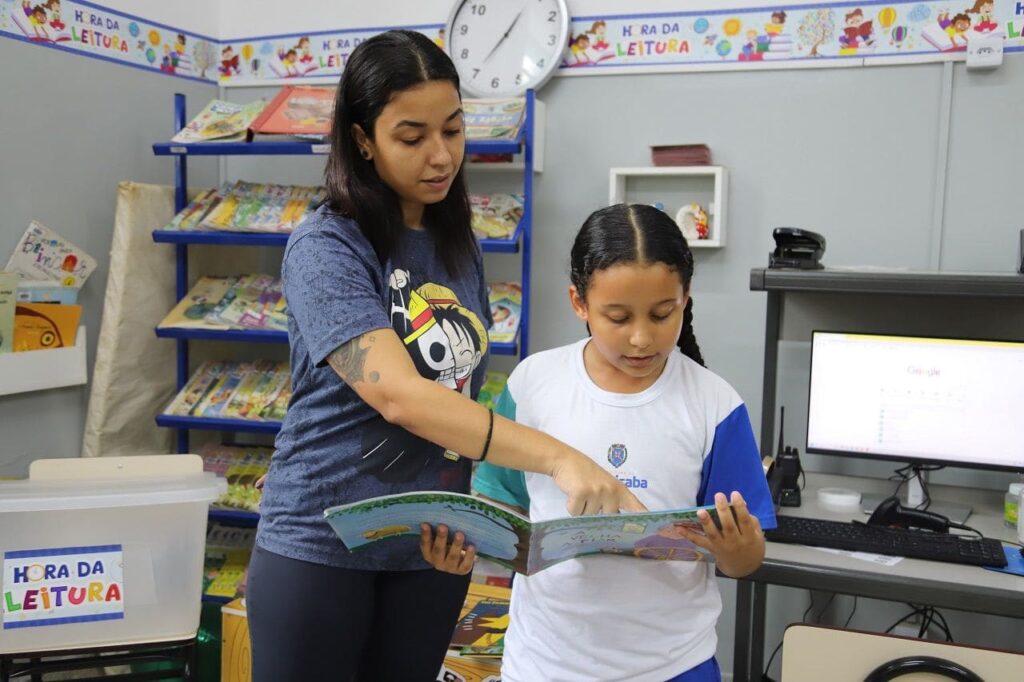 A imagem mostra uma cena em uma sala de aula ou biblioteca voltada para crianças. Uma mulher adulta está em pé ao lado de uma menina que parece ser aluna. A mulher, que tem cabelos escuros presos e tatuagens no braço, está vestindo uma camiseta azul escura com uma estampa de personagem e está apontando para um livro aberto que a menina segura. A menina, que usa um uniforme escolar branco com detalhes azuis, está concentrada na leitura do livro. Ao fundo, há estantes cheias de livros infantis coloridos, um computador com a tela aberta em uma página de busca do Google, e um relógio de parede. Na frente, há uma caixa transparente com a inscrição "HORA DA LEITURA", que também aparece repetida em uma faixa decorativa na parede, junto com desenhos de crianças lendo, reforçando o tema da leitura e educação. A iluminação é clara, criando um ambiente acolhedor e propício para o aprendizado. A imagem enfatiza a interação entre a professora e a aluna, destacando a importância do momento de leitura e da aprendizagem conjunta.