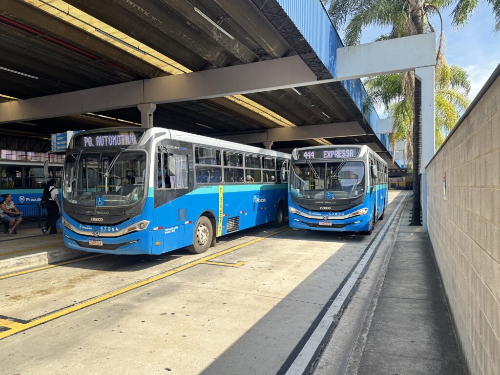 A imagem mostra um terminal de ônibus ao ar livre em Piracicaba, Brasil, em um dia ensolarado com céu azul. Dois ônibus modernos, pintados em azul e branco, estão estacionados lado a lado sob um grande teto metálico. O ônibus à esquerda exibe o número 57064 e tem o letreiro digital indicando o destino "PQ. AUTOMOTIVO". O ônibus à direita, número 57035, apresenta o letreiro "444 EXPRESSA". Ambos são da marca Mascarello, com chassis Iveco, fabricados em 2024. À esquerda da imagem, algumas pessoas aguardam o ônibus; uma está sentada em um banco e outra está em pé. O chão é de concreto com marcas amarelas que delimitam as faixas de circulação. À direita, há um muro branco e algumas palmeiras ao fundo, complementando o cenário. A luz do sol incide diretamente, criando sombras bem definidas sob a cobertura do terminal. A imagem transmite uma sensação de organização e modernidade no sistema de transporte público local.