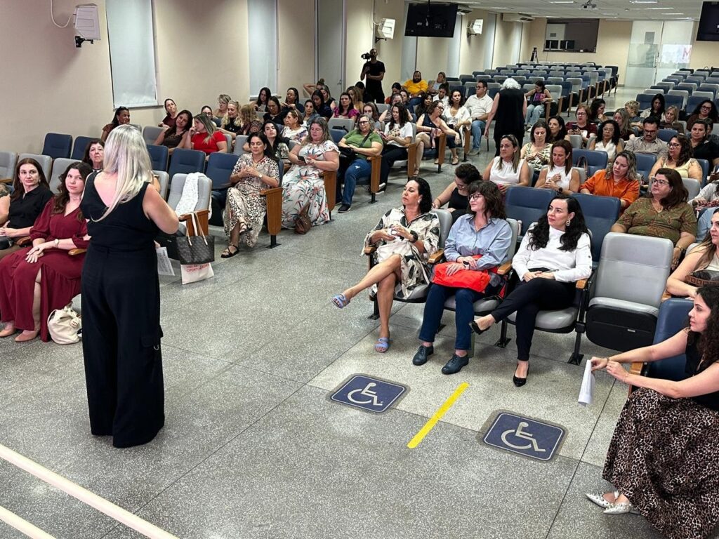 A imagem mostra uma sala de conferências ou auditório com várias pessoas sentadas, a maioria mulheres, ouvindo uma palestra ou apresentação. No centro da imagem, uma mulher está em pé, de costas para a câmera, provavelmente a palestrante, vestindo calças largas pretas e uma blusa preta sem mangas. As cadeiras são azuis e organizadas em filas, algumas ocupadas e outras vazias. No chão há marcas amarelas e símbolos azuis que indicam espaços reservados para cadeirantes. O ambiente é iluminado artificialmente, com um piso cinza e paredes claras. A atmosfera parece formal e focada na palestra. Algumas pessoas seguram papéis e bolsas estão apoiadas próximas às cadeiras. A imagem é capturada de um ângulo levemente elevado, mostrando a interação entre a palestrante e o público.