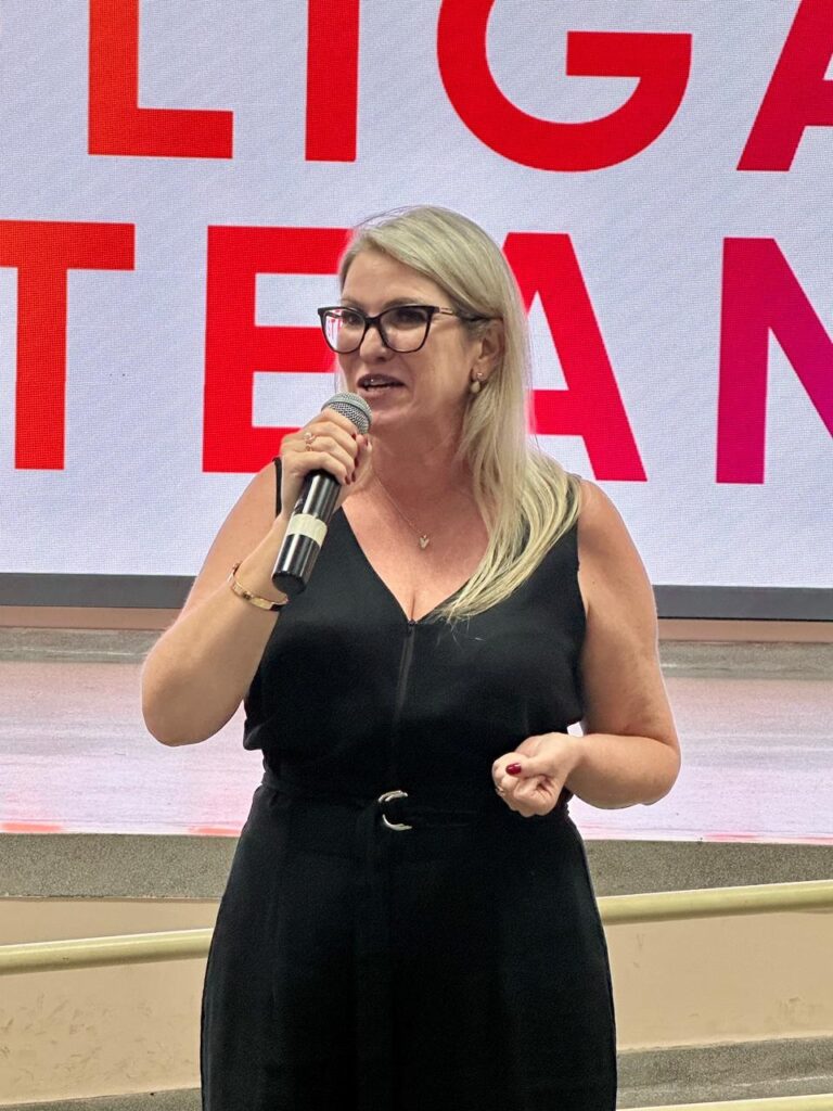 A imagem mostra uma mulher loira, usando óculos, falando ao microfone. Ela veste um vestido preto sem mangas, com um zíper na frente e um cinto com fivela prateada na cintura. A mulher também está usando um colar e pulseiras. O fundo apresenta uma tela grande com cores em tons de vermelho e rosa, exibindo texto que parece ser um logotipo ou slogan, embora o texto não esteja legível na imagem. A iluminação é clara, destacando a mulher e o fundo. A foto é um plano médio, capturando a mulher da cintura para cima, com uma perspectiva ao nível dos olhos. A cena sugere que ela está fazendo uma apresentação ou discurso em um evento.