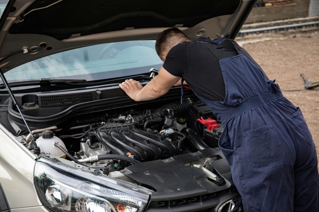 A imagem mostra um mecânico, um jovem de cabelo curto, vestindo uma camiseta preta e um macacão azul, inclinado sobre o compartimento do motor de um carro com o capô aberto. Ele está concentrado, com as mãos apoiadas na borda do motor, aparentemente inspecionando ou trabalhando no motor. O capô do carro está completamente aberto, sustentado por uma haste de apoio. O motor é moderno, com várias peças de plástico preto, fios e mangueiras visíveis. À direita, é possível ver o terminal vermelho da bateria. O carro parece ser um sedã moderno, com faróis e parte da grade frontal visíveis. O fundo está um pouco desfocado, mostrando um chão de cascalho e uma estrutura que pode ser uma cerca, sugerindo que o local é ao ar livre, possivelmente uma oficina ou garagem. A iluminação é natural, indicando que a foto foi tirada durante o dia, com luz difusa que evita sombras fortes. A composição destaca o mecânico e o motor do carro como foco principal, evidenciando o trabalho de manutenção automotiva.