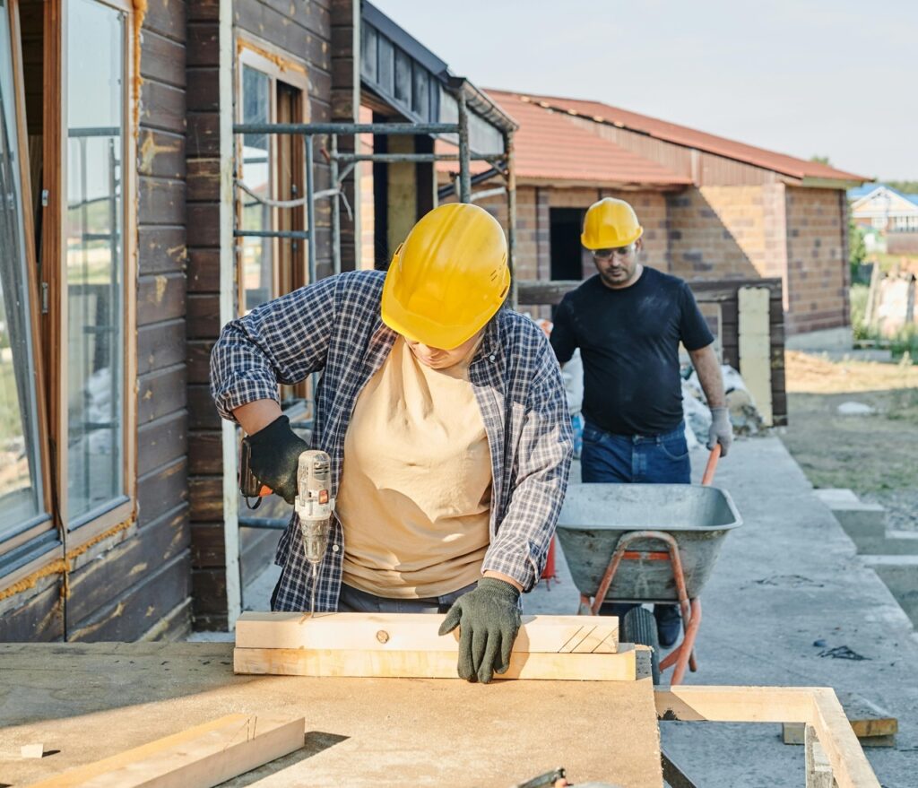 A imagem mostra dois trabalhadores da construção em um dia ensolarado em um canteiro de obras residencial. O foco principal é em um trabalhador no primeiro plano, usando um capacete amarelo, uma camisa xadrez sobre uma camiseta bege e luvas escuras. Ele está usando uma furadeira sem fio para fixar um parafuso em um pedaço de madeira que está apoiado em uma bancada de trabalho. No fundo, o segundo trabalhador, também com capacete amarelo, está empurrando uma carrinho de mão. Ele veste uma camiseta preta, jeans azuis e luvas, parecendo estar transportando materiais. Ao fundo, há uma casa de madeira parcialmente construída, com estrutura exposta e andaimes, além de uma casa de tijolos visível à distância, sugerindo um bairro em desenvolvimento. A iluminação é brilhante e natural, criando sombras marcantes, e a paleta de cores é dominada por tons terrosos de madeira e tijolo, contrastando com o amarelo vibrante dos capacetes e o azul do céu. A composição enfatiza a ação do trabalhador com a furadeira, enquanto o fundo contextualiza o ambiente de construção. A perspectiva é ao nível dos olhos, capturando a cena de forma direta.