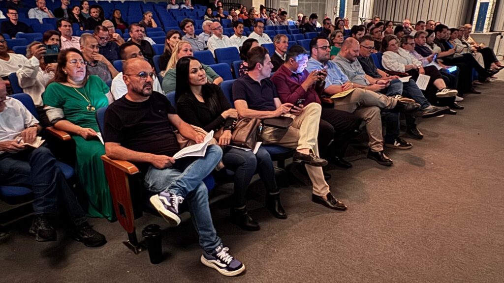 A imagem mostra uma plateia sentada em cadeiras azuis estilo teatro, com braços e bases de madeira. As pessoas estão voltadas ligeiramente para a direita, aparentemente assistindo a uma apresentação ou palestra. A iluminação é artificial e cria um ambiente acolhedor. Na frente, há um homem de camiseta preta e jeans segurando um papel, ao lado de uma mulher vestindo um vestido verde vibrante e um colar dourado, também com um papel na mão. Ao lado dela, outro homem de camiseta preta e óculos olha para a direita. Ao fundo, diversas outras pessoas estão sentadas, algumas olhando para a frente, outras segurando celulares, uma delas com o rosto iluminado pela tela do aparelho. A atmosfera é de concentração e atenção, típica de um evento cultural ou educacional. O piso é de carpete marrom escuro e a parede ao fundo tem painéis verticais.