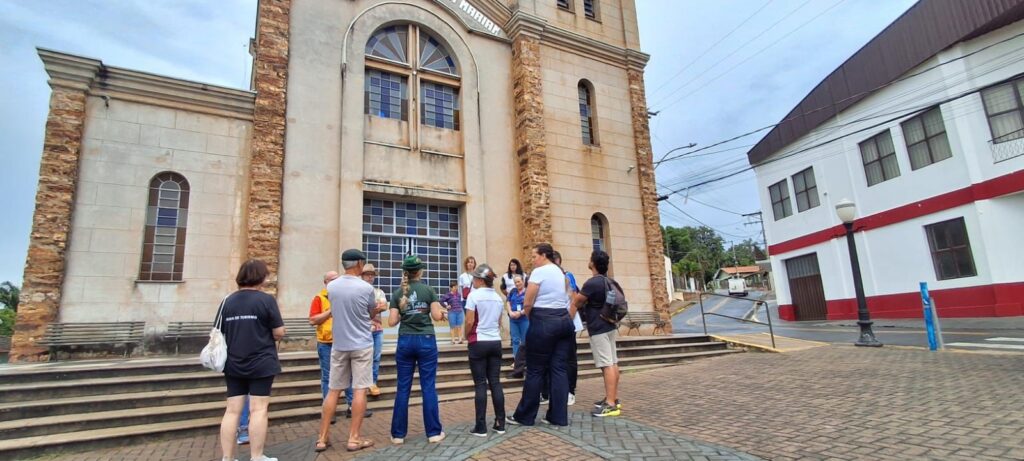 A imagem mostra um grupo de pessoas reunidas em frente a uma igreja de pedra com arquitetura antiga. A igreja tem uma fachada feita de blocos de pedra clara com cantos em pedras de tom marrom-avermelhado. A porta principal da igreja está fechada por um portão de metal, e acima da porta é possível ver parcialmente a palavra "MARIAN". As pessoas parecem estar participando de um passeio guiado, pois uma mulher está usando uma camiseta preta com a inscrição "GUIA DE TURISMO" nas costas. O grupo está distribuído na calçada e nos degraus em frente à igreja, ouvindo a guia que fala para eles. A maioria está vestida de forma casual, com camisetas, shorts e calças, e alguns usam chapéus. Ao lado direito da igreja há um prédio branco com detalhes em vermelho, e uma luminária de rua está visível. O céu está nublado, indicando um dia com pouca luz solar direta. O ambiente transmite a sensação de um passeio turístico cultural, possivelmente em uma cidade histórica.