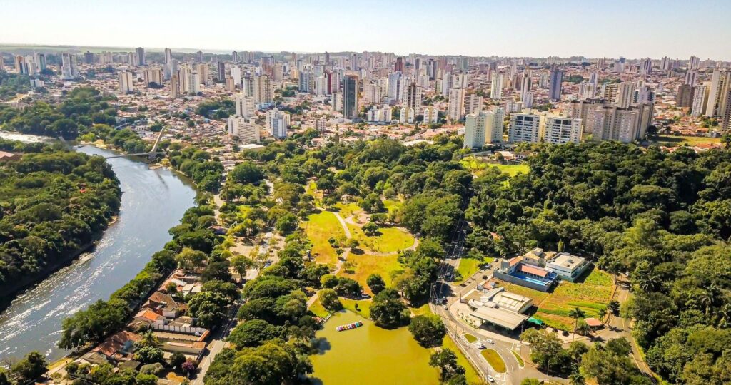 A imagem mostra uma vista aérea de uma grande cidade onde o contraste entre natureza e urbanização se destaca. Em primeiro plano, um rio sinuoso atravessa a paisagem, ladeado por áreas verdes densas. Ao lado dele, há um amplo parque com campos gramados, caminhos para caminhada e grupos de árvores, além de um lago menor com barcos coloridos. Esse espaço verde é cercado por edifícios residenciais e comerciais, e ao fundo surge uma concentração de arranha-céus que compõem o horizonte urbano.