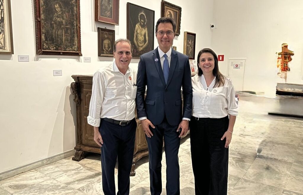 Na imagem aparecem ao centro um homem de óculos de terno azul escuro, olhando para a câmera. Ao seu lado direito aparece um senho de camisa clara e calça escura, sorrindo e ao seu lado esquerdo, aparece uma mulher que está sorrindo, vestindo camisa clara e calça escura. Estão em uma sala com diversos quadros na parede.