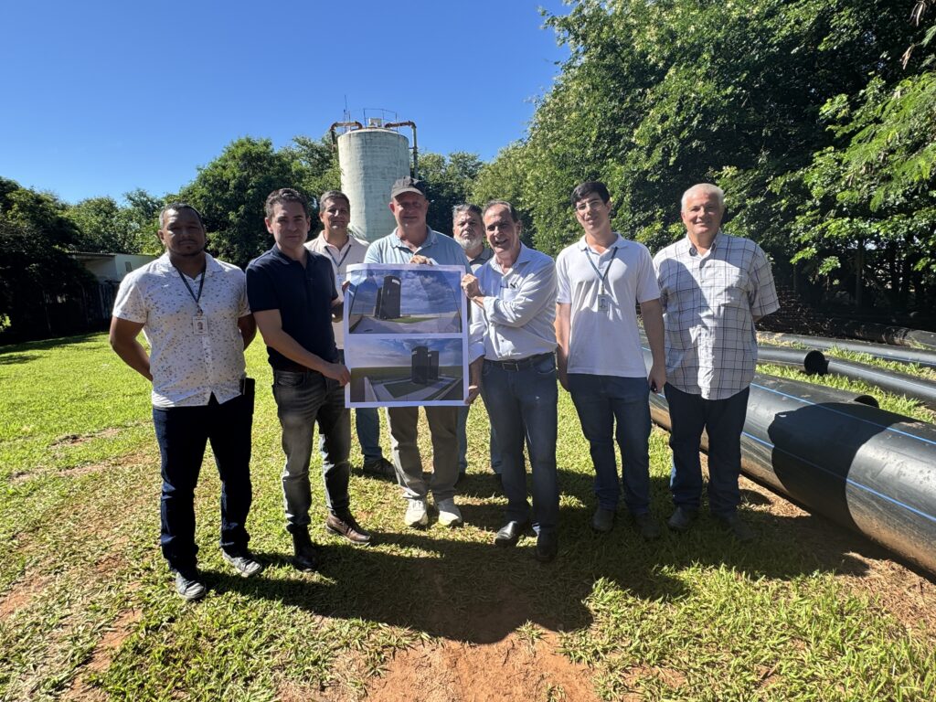 A imagem mostra um grupo de oito homens em um ambiente externo, em uma área gramada com árvores ao fundo. Eles estão posando para a foto, com a maioria olhando para a câmera. Dois dos homens no centro seguram um grande cartaz que exibe imagens renderizadas de estruturas industriais, provavelmente tanques de armazenamento ou silos, mostrados de ângulos diferentes com céu azul ao fundo. À direita do grupo, há vários tubos grandes e pretos sobre a grama, possivelmente para uso em sistemas de água ou esgoto. Ao fundo, é possível ver uma torre de água alta, cilíndrica, com uma plataforma no topo, rodeada por árvores verdes, sugerindo que a foto foi tirada em um dia ensolarado. Os homens estão vestidos de forma casual, com camisas de manga curta ou pólos e calças compridas. Um deles usa boné. O cenário aparenta ser uma visita técnica ou apresentação de um projeto relacionado à infraestrutura ou saneamento.