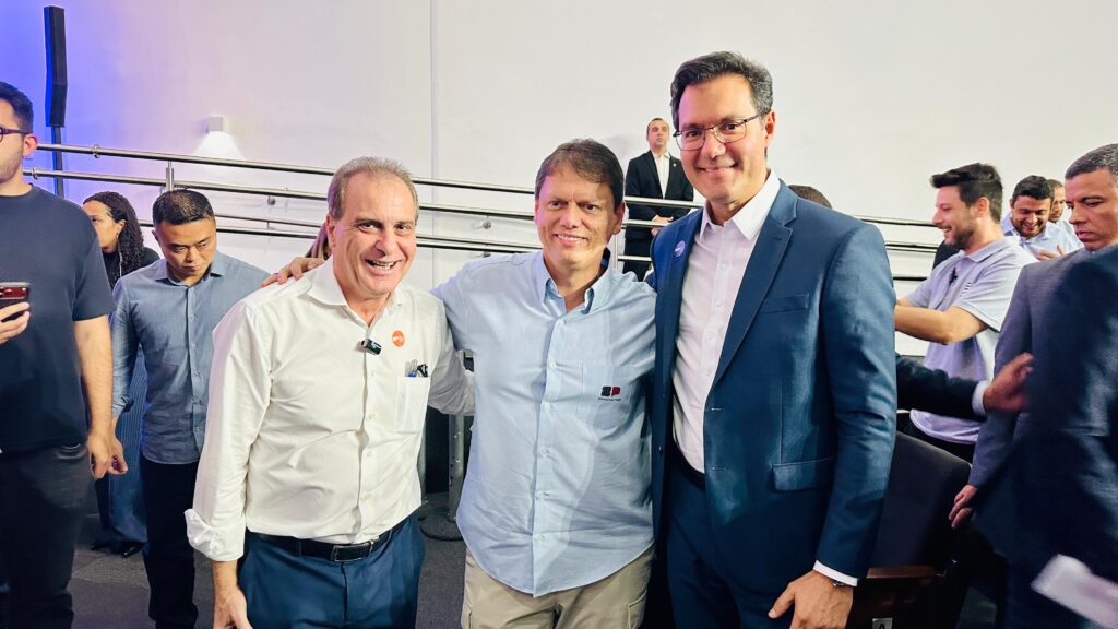 Na imagem aparecem três homens, tendo ao centro um homem sorridente, tendo ao lado direito um senhor sorridente de camisa clara e calça azul e no lado esquerdo um rapaz de óculos, camisa branca e terno azul escuro.