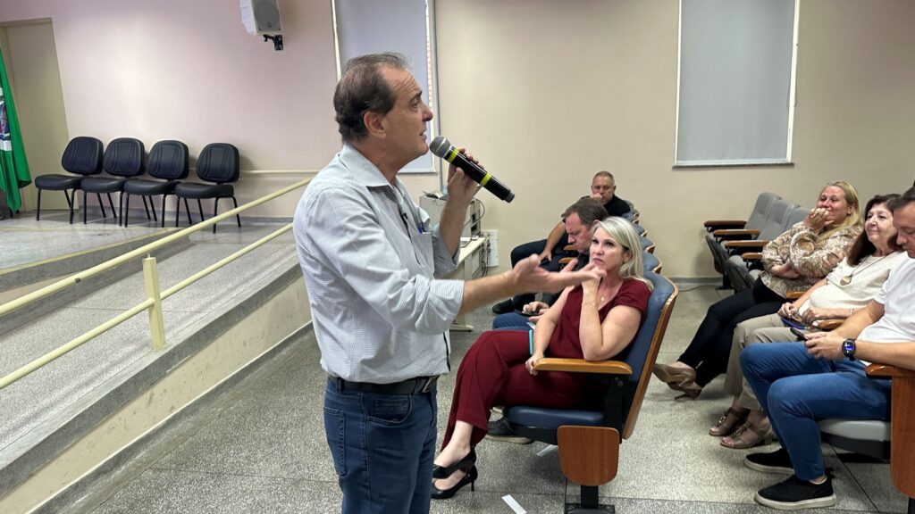 A imagem mostra um homem falando em público dentro de um auditório. Ele está em pé, segurando um microfone sem fio na mão direita e gesticulando com a mão esquerda. Ele veste uma camisa clara com estampa e calça jeans. Ao fundo, há um palco elevado com quatro cadeiras pretas e uma bandeira verde ao lado. O palco tem uma escada com corrimão metálico. Sentadas em cadeiras de auditório, algumas pessoas assistem à apresentação; entre elas, uma mulher de macacão vermelho na primeira fila, olhando atentamente para o palestrante. O ambiente é iluminado por luzes fluorescentes, com paredes claras e piso de azulejo cinza. A atmosfera parece formal, possivelmente um evento, palestra ou reunião.