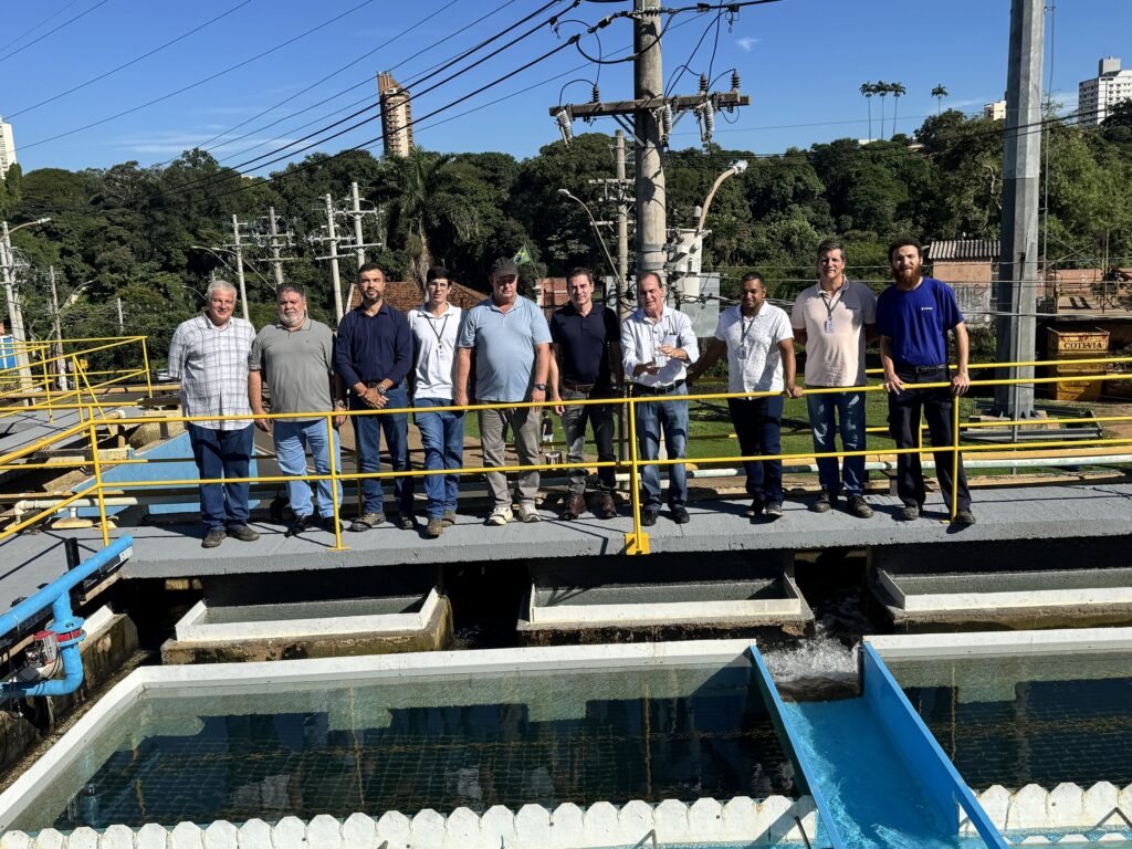 A imagem mostra um grupo de homens em uma passarela acima de uma estação de tratamento de água. Eles estão observando as estruturas da estação, que incluem canais de água com grades amarelas de proteção nas laterais. O ambiente é industrial, com tubulações, tanques e edifícios ao fundo. O céu está claro e azul, indicando um dia ensolarado. Ao redor da estação há vegetação e algumas construções urbanas. Os homens parecem estar em uma visita técnica ou inspeção, vestidos de forma casual e alguns carregando mochilas. A cena transmite uma sensação de trabalho e monitoramento em um ambiente de infraestrutura pública.