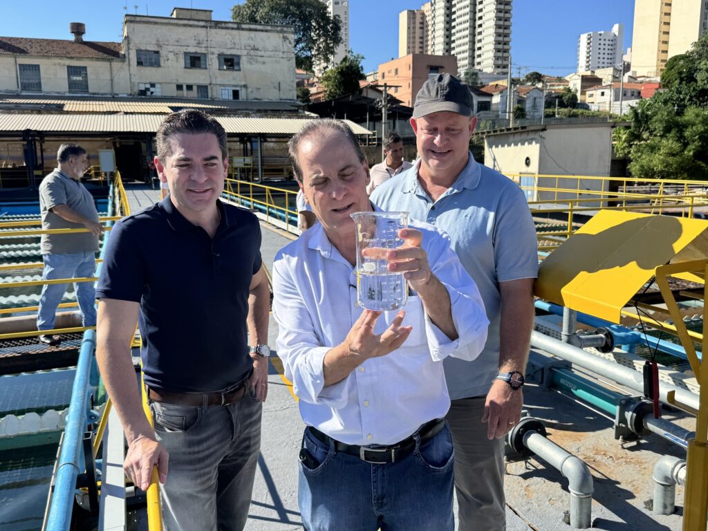 A imagem mostra três homens em uma plataforma industrial, provavelmente em uma estação de tratamento de água, sob um céu azul claro. O homem no centro, vestido com uma camisa azul clara e jeans, segura um béquer de vidro com líquido transparente, observando-o atentamente. Ele parece estar explicando algo, com uma expressão focada e um leve sorriso. À esquerda dele, outro homem, usando uma camisa polo azul escura e jeans cinza, sorri confiante olhando para a câmera. À direita do homem com o béquer, há um terceiro homem com uma camisa polo azul clara, calça escura e boné, também olhando para o centro com um leve sorriso. Ao fundo, um quarto homem caminha afastado, perto de uma estrutura industrial com fachadas desgastadas e prédios altos modernos ao longe. A plataforma tem corrimãos amarelos e equipamentos industriais visíveis, como tubulações. A iluminação é forte, indicando que a foto foi tirada durante o dia, com sombras bem definidas. No béquer que o homem segura, está escrito "Satelit 1000 mL" e "NETZSCH", indicando a marca e a capacidade do recipiente. A cena transmite uma atividade de inspeção ou análise da qualidade da água, com os homens aparentando estar trabalhando ou realizando uma verificação técnica no local.