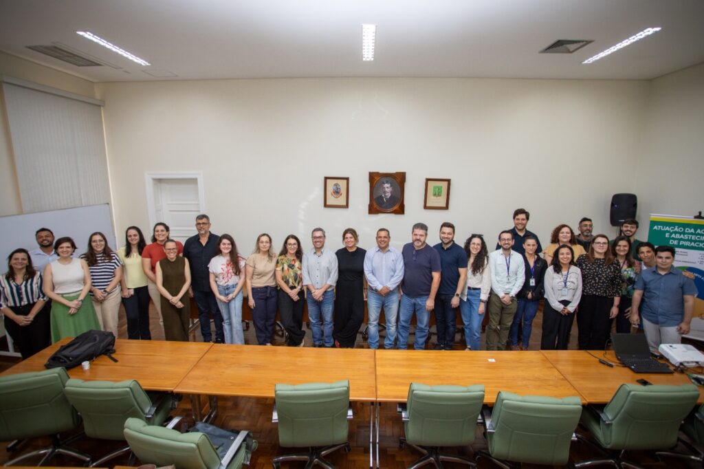 A imagem mostra um grupo de 31 pessoas, sendo 15 homens e 16 mulheres, posando para uma foto em um ambiente que parece ser uma sala de conferências ou escritório. A maioria está em pé, formando uma fila atrás de uma longa mesa de madeira, enquanto alguns estão sentados em cadeiras verdes à frente da mesa. O ambiente é bem iluminado com luz fluorescente, com paredes claras e um teto branco. Ao fundo, há um quadro branco à esquerda, duas imagens ou quadros pendurados na parede e um alto-falante à direita. Sobre a mesa, há um laptop e um projetor, indicando que pode ter ocorrido uma apresentação ou reunião. A maioria das pessoas está vestida de forma casual e profissional, com camisas, blusas e calças. As expressões faciais são em sua maioria neutras ou com sorrisos leves, sugerindo uma foto formal de grupo. Na lateral direita da imagem, há um cartaz com o texto "ATUAÇÃO DA ÁGUA E ABASTECIMENTO DE PIRACICABA", indicando que o grupo pode estar relacionado a um projeto ou trabalho envolvendo a água e o abastecimento na cidade de Piracicaba. A composição da foto é centralizada no grupo, com a mesa e as cadeiras criando profundidade na imagem.