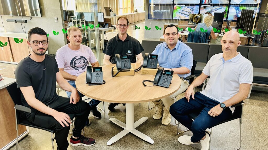 A imagem mostra cinco homens sentados ao redor de uma mesa redonda de madeira clara em um ambiente de escritório moderno. Eles estão posicionados de forma simétrica, olhando diretamente para a câmera, com expressões neutras a amigáveis. Na mesa, há quatro telefones VoIP pretos, dois ainda em suas embalagens de papelão. Os homens vestem roupas casuais e formais: o primeiro à esquerda usa camiseta preta e óculos; o segundo, camiseta rosa com estampa; o do centro, polo preta e óculos; o quarto, camisa azul clara e óculos; e o último, polo branca com calça escura. Ao fundo, há divisórias de vidro com adesivos de folhas verdes, computadores e uma grande parede de vídeo exibindo imagens de câmeras ou dados. O ambiente é bem iluminado, com piso cinza, possivelmente de concreto polido ou azulejo. A composição é uma foto de média distância, capturando os homens da cintura para cima. No canto da imagem, há textos que indicam "Centro de Inovação Prefeitura de Joinville", além de mensagens de erro relacionadas a rede.