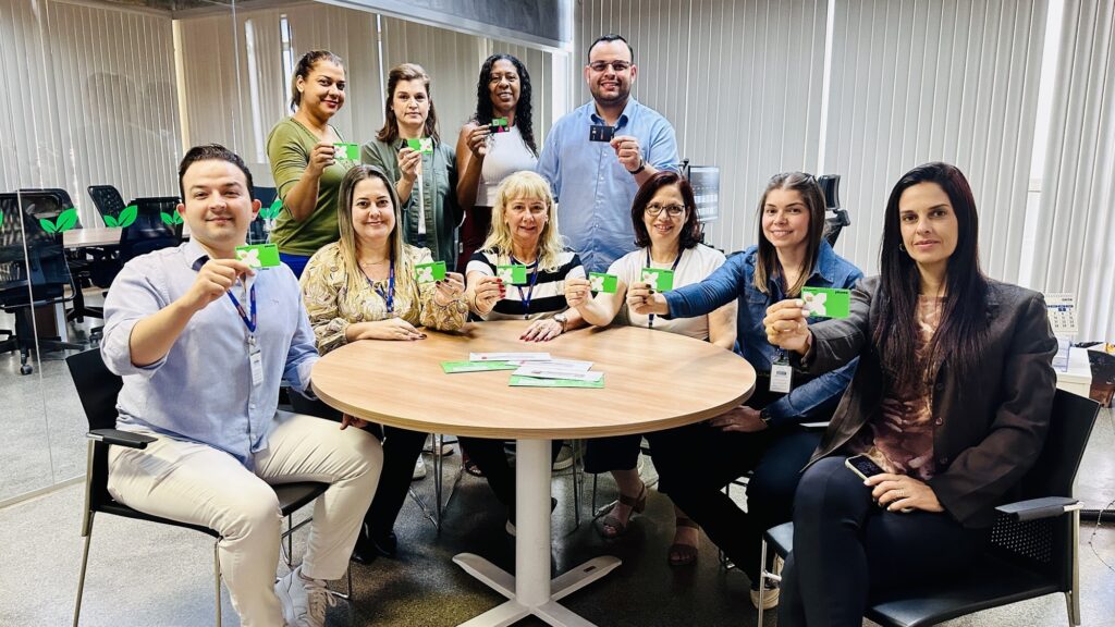 A imagem mostra um grupo de dez pessoas, sendo oito mulheres e dois homens, reunidas em torno de uma mesa redonda de madeira em um ambiente de escritório. A maioria está segurando pequenos cartões verdes com um "X" branco e a palavra "phone" impressa. Eles estão vestidos de forma casual e parecem estar em um momento descontraído e positivo, sorrindo para a câmera. O ambiente ao fundo inclui cadeiras de escritório, mesas e janelas com persianas, indicando que é um espaço de trabalho. A iluminação é clara e uniforme, e a foto transmite um clima profissional, porém amigável e colaborativo.
