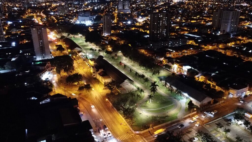 A imagem mostra uma vista aérea noturna de um centro urbano, com uma praça ou parque público bem iluminado no centro. O parque possui um caminho em forma de laço, áreas verdes e várias palmeiras, todas iluminadas por refletores que destacam o espaço em meio à escuridão da noite. Ao redor do parque, há ruas com veículos em movimento, evidenciados pelo efeito de luzes em movimento, e algumas faixas de pedestres visíveis. Vários prédios altos, possivelmente residenciais ou comerciais, cercam a área, criando um contraste entre as construções mais altas e as casas de menor altura espalhadas pela cidade. A iluminação urbana é composta por luzes amarelas dos postes e luzes brancas dos edifícios, conferindo um ambiente acolhedor e vibrante, apesar do horário noturno. O cenário sugere uma cidade moderna, com planejamento urbano que valoriza espaços verdes no meio da malha urbana.