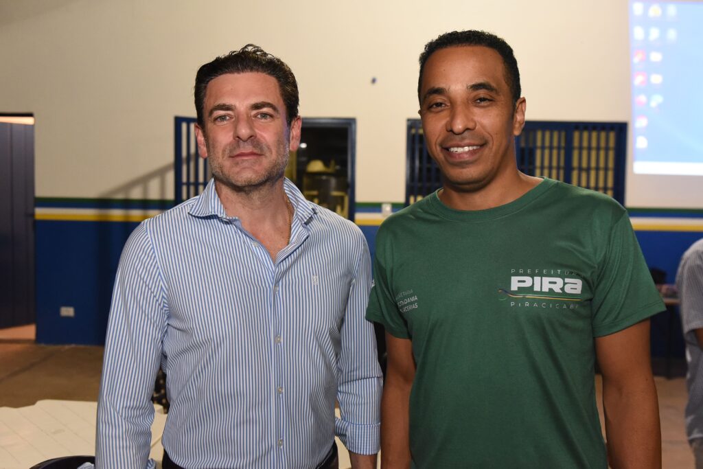 A imagem mostra dois homens posando para uma foto em um ambiente interno. O homem à esquerda veste uma camisa listrada azul e branca e usa uma corrente dourada no pescoço. Ele tem cabelo escuro, pele clara e uma expressão séria. O homem à direita usa uma camiseta verde com um desenho branco e sorri amplamente. Ele tem cabelo escuro e encaracolado e pele mais escura. Ao fundo, há uma parede clara com detalhes em azul e amarelo, além de uma tela de projeção exibindo alguns ícones coloridos. A iluminação é artificial e uniforme, destacando bem os rostos dos homens. A foto parece ter sido tirada em um evento ou reunião formal ou semi-formal. No canto da imagem, é possível ler parte de um banner ou cartaz com as palavras "PREFEITURA", "PIRACICABA" e "SECRETARIA CIDADANIA".