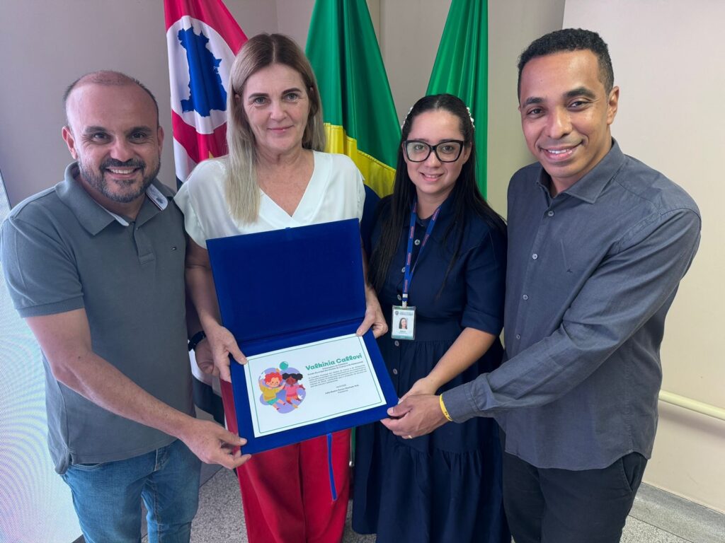 A imagem mostra quatro pessoas, duas mulheres e dois homens, posando para uma foto enquanto seguram um certificado ou placa azul. Eles estão sorrindo e parecem estar em um ambiente oficial, possivelmente uma cerimônia de premiação ou reconhecimento. Ao fundo, há bandeiras que indicam um contexto institucional, provavelmente relacionado ao município de Piracicaba, conforme o texto no certificado. O certificado que eles seguram tem uma ilustração colorida na parte inferior, mostrando crianças ao redor de um globo terrestre, simbolizando educação e cultura. O texto no certificado parabeniza Valdikiria Callovi pela sua contribuição para o desenvolvimento da educação e cultura no município de Piracicaba, datado de 25 de setembro de 2023, assinado pelo prefeito municipal Adilson Roberto Furlan. Os quatro indivíduos estão vestidos de forma casual a semi-formal, com uma mulher no centro usando uma blusa branca e calça vermelha, e os demais com roupas em tons neutros. A atmosfera é de celebração e reconhecimento público.