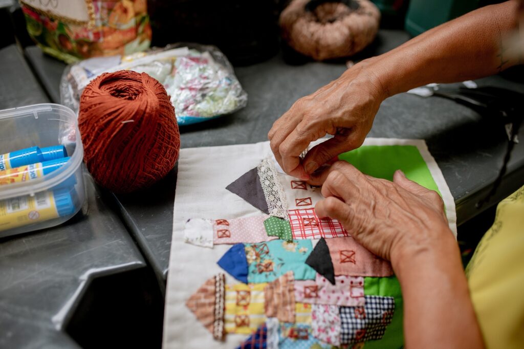 A imagem mostra um close das mãos de uma pessoa idosa trabalhando em um projeto de costura ou patchwork. As mãos estão cuidadosamente alinhando ou costurando pequenos pedaços de tecido colorido e estampado, que formam um padrão detalhado, parecido com pequenas casinhas com telhados. Ao lado do tecido, há um novelo grande de linha ou lã em tom ferrugem. Também é possível ver um recipiente de plástico transparente contendo dois bastões de cola. O ambiente sugere um espaço de artesanato, com outros materiais como sacos de tecido ao fundo e uma superfície escura onde a pessoa trabalha. A iluminação destaca as texturas das mãos, dos tecidos e do novelo, transmitindo uma sensação de cuidado e dedicação ao trabalho manual.