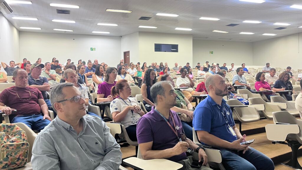 A imagem mostra uma plateia sentada em um auditório, provavelmente durante uma palestra, conferência ou curso. As pessoas estão organizadas em fileiras de cadeiras com pequenas mesas acopladas, dispostas em degraus, o que facilita a visão para todos. A iluminação é clara e uniforme, vinda de luminárias no teto, criando um ambiente bem iluminado. No primeiro plano, há alguns participantes em destaque: um homem de camisa azul clara com óculos, outro de camiseta vermelha escura com óculos pendurados no colarinho, um homem de camisa polo roxa segurando óculos e celular, e outro de camisa polo azul com crachá, também com celular na mão. A maioria das pessoas está atenta, olhando para a frente, possivelmente para um palestrante ou tela de apresentação, enquanto algumas olham para baixo, talvez consultando anotações ou celulares. O público é diversificado em termos de idade e gênero, com vestimentas que variam entre casual e casual social, condizentes com um ambiente profissional ou educacional. Ao fundo, há mais pessoas sentadas, preenchendo o auditório, que tem paredes claras e um teto com ventilação visível. Há também uma sinalização de saída de emergência iluminada. Algumas pessoas usam crachás e lanyards, e é possível ver a marca "SEBRAE" em algumas camisas e crachás, indicando que o evento pode estar relacionado a essa instituição. Um detalhe colorido é uma mochila com estampa floral colocada em uma das cadeiras próximas ao primeiro plano. No geral, a imagem transmite a atmosfera de um evento formal de aprendizado ou capacitação, com um público engajado e atento.