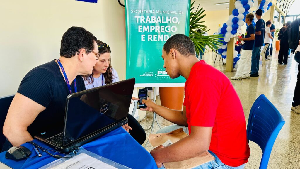 A imagem mostra uma cena em um ambiente interno, possivelmente uma prefeitura ou centro de atendimento público, onde ocorre um serviço de atendimento ao cidadão. No centro da imagem, um jovem vestido com uma camiseta vermelha está sentado em frente a uma mesa azul, mostrando a tela do seu celular para duas mulheres que estão do outro lado da mesa. Uma das mulheres tem cabelo curto e usa óculos, vestindo uma blusa preta, enquanto a outra tem cabelo longo e está com uma blusa branca. Sobre a mesa, há um notebook da marca Dell e alguns papéis. Atrás das mulheres, há um banner verde com o logo da Prefeitura de Piracicaba e o texto "SECRETARIA MUNICIPAL DE TRABALHO, EMPREGO E RENDA" e o slogan "FAZENDO O QUE PRECISA SER FEITO". Ao fundo, outras pessoas aguardam atendimento, indicando que se trata de um local de serviços públicos, provavelmente relacionado a emprego e renda. A iluminação é boa, com luz natural vindo das janelas e luz artificial do ambiente, criando um cenário claro e acolhedor. A interação entre as pessoas sugere um atendimento personalizado, onde o jovem está recebendo ajuda ou esclarecimentos sobre algum serviço.