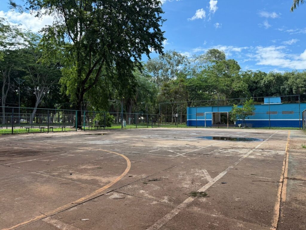 A imagem mostra uma quadra de basquete ao ar livre, com o piso de concreto apresentando marcas de desgaste e algumas poças de água, indicando que pode ter chovido recentemente. As linhas da quadra, pintadas em branco e amarelo, estão um pouco desbotadas. Ao fundo, há um prédio azul com detalhes em branco, possivelmente uma escola ou centro esportivo, com janelas e uma estrutura que parece uma trave, sugerindo que o local pode ser usado para diferentes esportes. A quadra está cercada por uma grade metálica, e ao redor há árvores e vegetação, conferindo um ambiente aberto e natural. O céu está azul com algumas nuvens brancas espalhadas, e a luz do sol ilumina a cena, indicando que é dia. A quadra aparenta estar um pouco desgastada, com rachaduras e sujeira visíveis no chão.