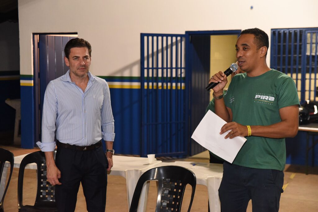A imagem mostra dois homens em um ambiente interno, possivelmente durante um evento ou reunião. O homem à esquerda está vestido com uma camisa de manga longa listrada azul e branca, calça escura e usa um relógio no pulso esquerdo. Ele tem cabelo escuro e está olhando levemente para a direita, com uma expressão neutra. O homem à direita veste uma camiseta verde com o logo da "PREFEITURA PIRA PIRACICABA" estampado na parte frontal. Ele segura um microfone na mão direita e um papel branco na mão esquerda, parecendo estar falando ou apresentando algo. Também usa um relógio e uma pulseira amarela. Ao fundo, há uma mesa branca com cadeiras, um portão ou grade azul metálica e uma parede com faixas coloridas (amarela, verde e azul). Sobre a mesa, é possível ver alguns copos plásticos. A iluminação é artificial, criando sombras no ambiente. A foto é capturada na altura dos olhos, focando nos dois homens e sua interação.
