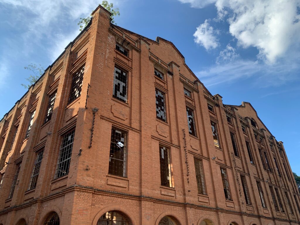 A imagem mostra um prédio industrial, construído em tijolos vermelhos, visto de um ângulo baixo que destaca sua altura imponente. O edifício tem várias janelas altas e estreitas, muitas delas com vidros quebrados ou ausentes. No topo e em algumas partes superiores do prédio, há vegetação crescendo, como pequenas árvores e ervas daninhas, indicando que a construção está abandonada há bastante tempo. O céu está claro, com algumas nuvens finas, e a luz do sol incide diretamente sobre a fachada, realçando a textura dos tijolos e criando sombras marcantes. A atmosfera geral transmite um sentimento de abandono e passagem do tempo.