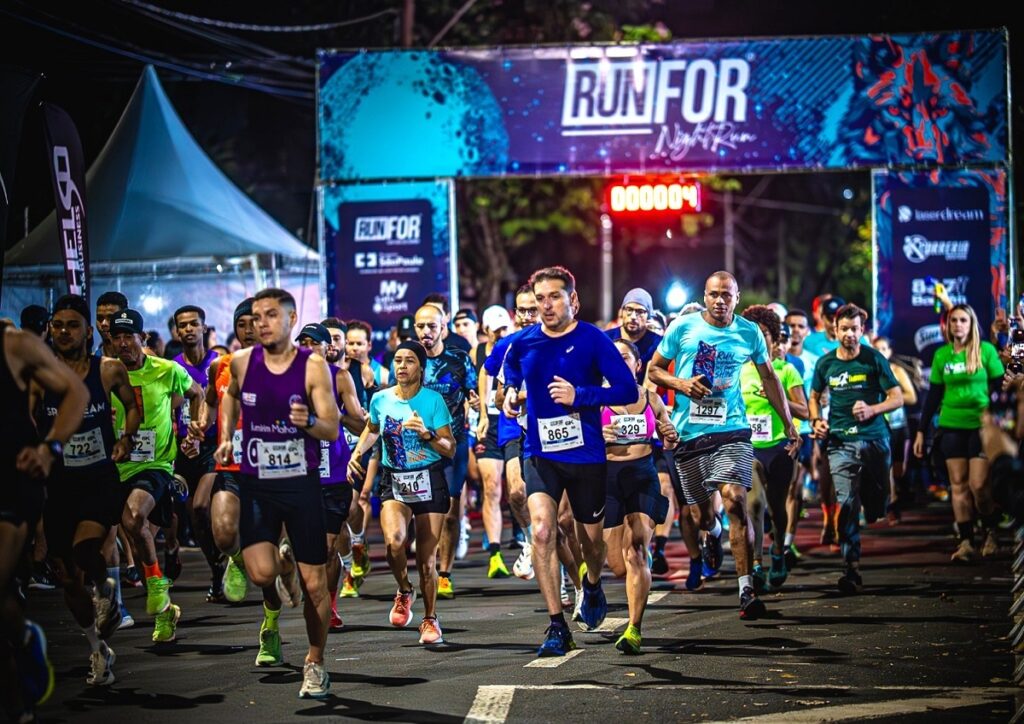 A imagem mostra o momento de largada de uma corrida noturna, intitulada "RUN FOR Night Run". Um grande grupo de corredores está passando por baixo de um arco inflável azul, que marca o ponto de largada ou chegada. O relógio digital no arco indica o tempo "000004", sugerindo que a prova acabou de começar. Os corredores estão vestidos com roupas esportivas coloridas, predominando tons de roxo, azul, verde-limão e turquesa. Entre eles, destacam-se alguns atletas com números de peito visíveis, como 814, 865, 1297, 210 e 329. Eles parecem concentrados e determinados, correndo em ritmo acelerado. Ao fundo, é possível ver uma tenda branca e algumas estruturas metálicas usadas para organizar o evento. A cena ocorre à noite, iluminada por luzes artificiais que criam contrastes fortes e destacam os corredores em primeiro plano. A atmosfera é de energia e movimento, típica de uma competição esportiva organizada em ambiente urbano.