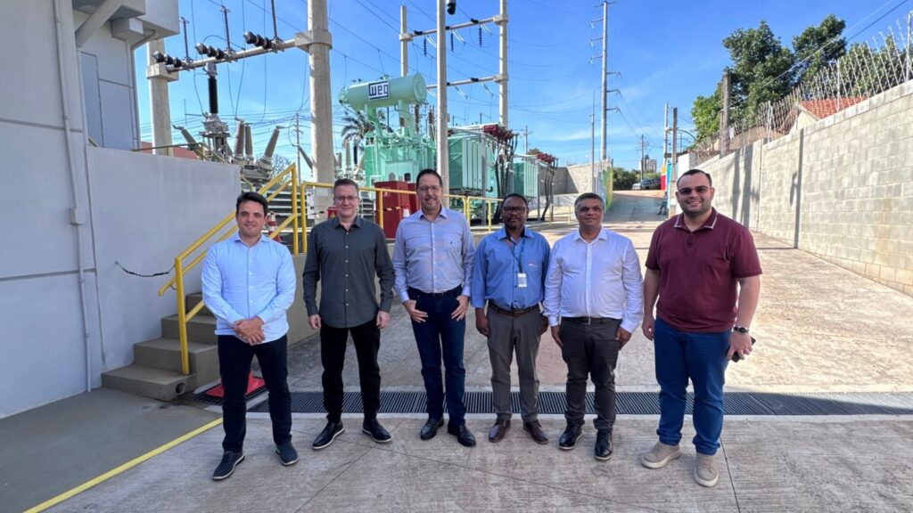 A imagem mostra seis homens em pé, alinhados, em um ambiente industrial ao ar livre, provavelmente uma subestação elétrica. Ao fundo, há equipamentos elétricos como transformadores, postes e cabos, além de uma construção com telhado vermelho e algumas árvores. O chão é pavimentado e há uma parede de concreto à direita da imagem. Os homens vestem roupas casuais e sociais, com camisas de manga curta e comprida, e parecem estar posando para a foto. O céu está claro e azul, indicando que a foto foi tirada durante o dia. A atmosfera geral sugere uma inspeção ou visita técnica ao local.