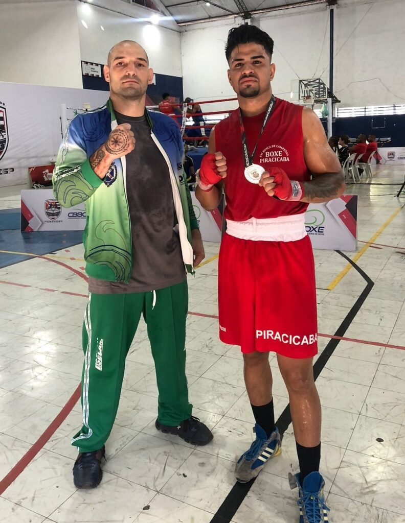 A imagem mostra dois homens em um ambiente de ginásio esportivo, provavelmente uma academia de boxe. O homem à esquerda, que parece ser o treinador, é careca, tem tatuagens nas mãos e veste uma camiseta cinza com uma jaqueta azul e verde por cima. Ele está com os punhos cerrados, numa pose de determinação. O homem à direita é um boxeador jovem, vestindo um uniforme vermelho com as inscrições "BOXE PIRACICABA" e "PIRACICABA" nas roupas. Ele também está com os punhos cerrados e usa uma medalha pendurada no pescoço, indicando que participou de uma competição. Ao fundo, vê-se um ringue de boxe, algumas pessoas e acessórios esportivos, além de banners com inscrições como "FEBOXESP", "CBBC" e "FEDERAÇÃO DE BOXE". O ambiente é iluminado por luz natural que entra pelas janelas superiores. A imagem transmite um clima de foco, esforço e conquista no esporte do boxe.